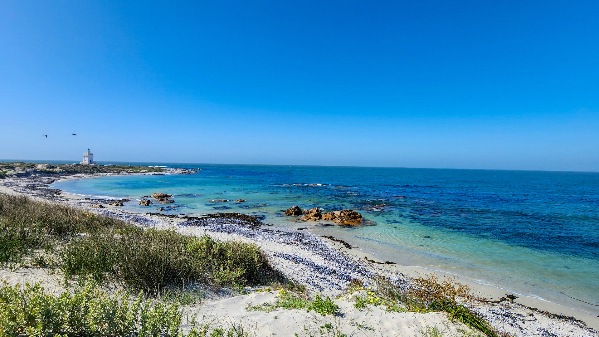 A quiet West Coast bay with turquoise water, white sand, and a distant lighthouse
